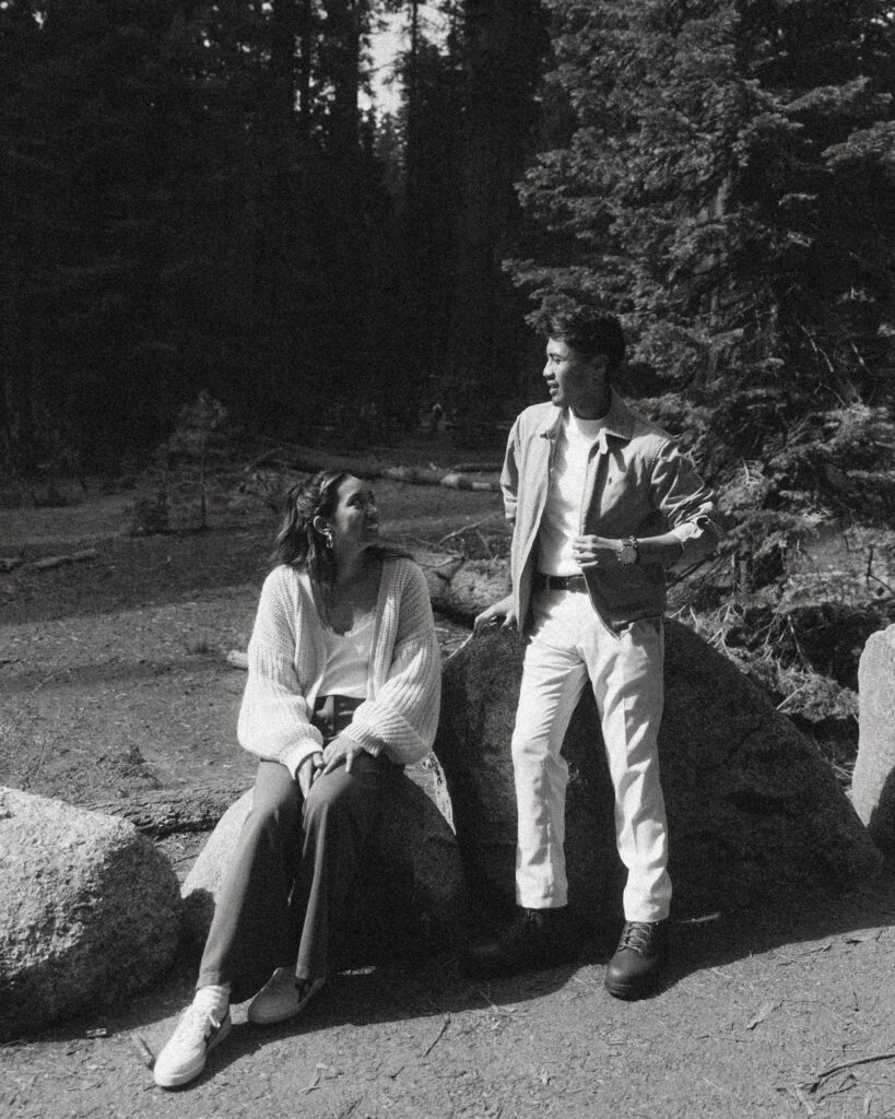 a black and white portrait of a couple smiling together while sitting on rocks an fallen trees