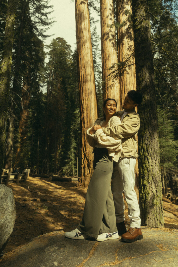 a man hugs his fiancee from behind as she smiles at him while they stand on a rock at sequoia national park