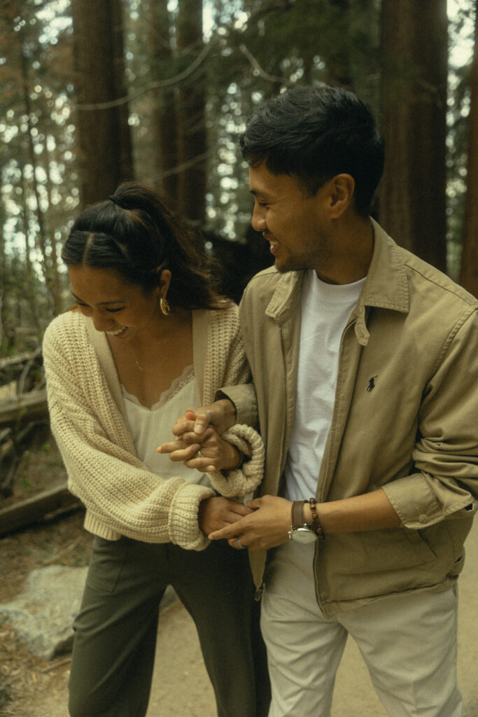 a couple laughs during their sequoia national park engagement photos while holding hands on a dirt path