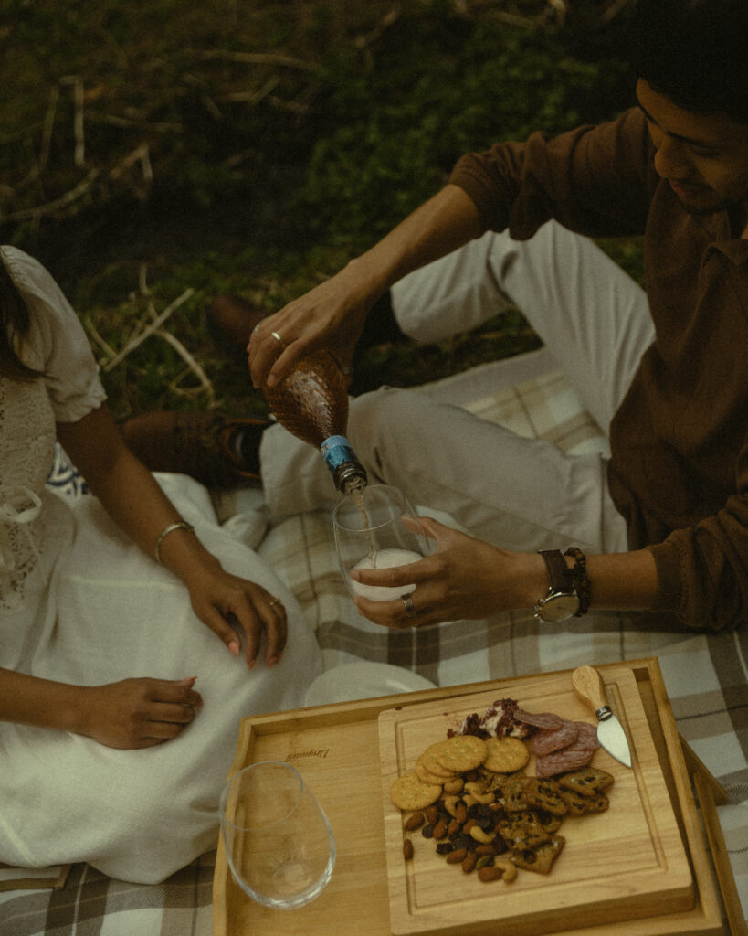 a couple shares snacks on a picnic blanket as he pours a bubbly drink into a glass in this cropped photo