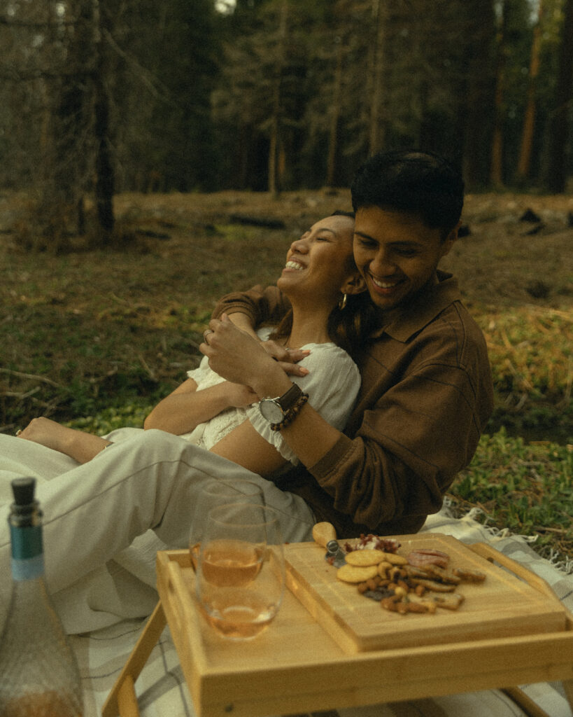 a couple laughs together while they share a picnic in the forest