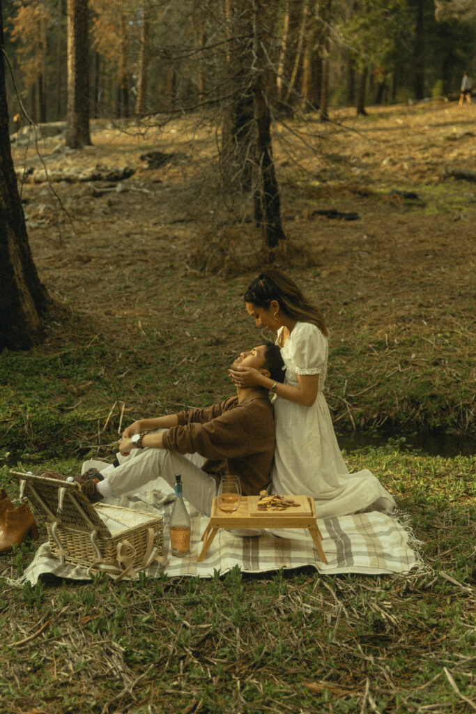 a woman kneels behind her fiancee on a plaid picnic blanket in the forest as he leans his head back to look up at her