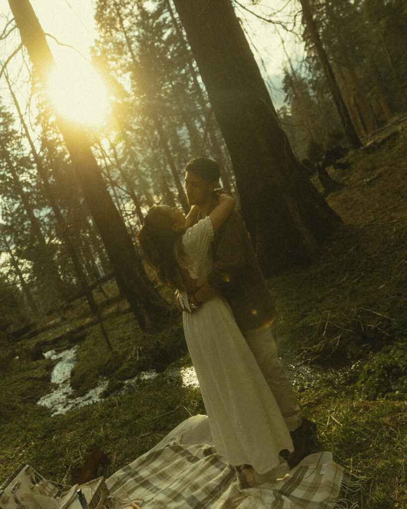 sequoia national park engagement photos where the couple dances together next to a picnic blanket placed by a stream with the light of the sun filtering through the trees
