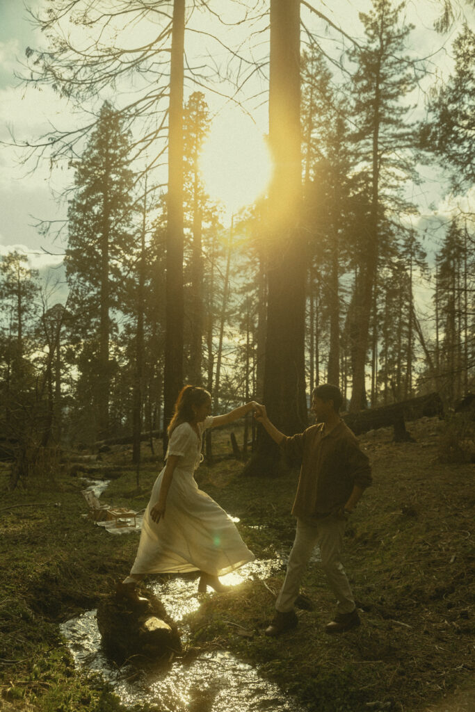 a man holds his hand to help his fiancee across a stream in the forest