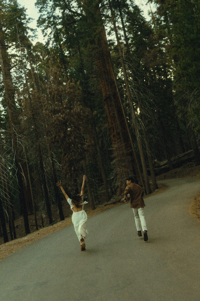 sequoia national park engagement photos where the couple runs excitedly down a paved road toward tall skinny trees