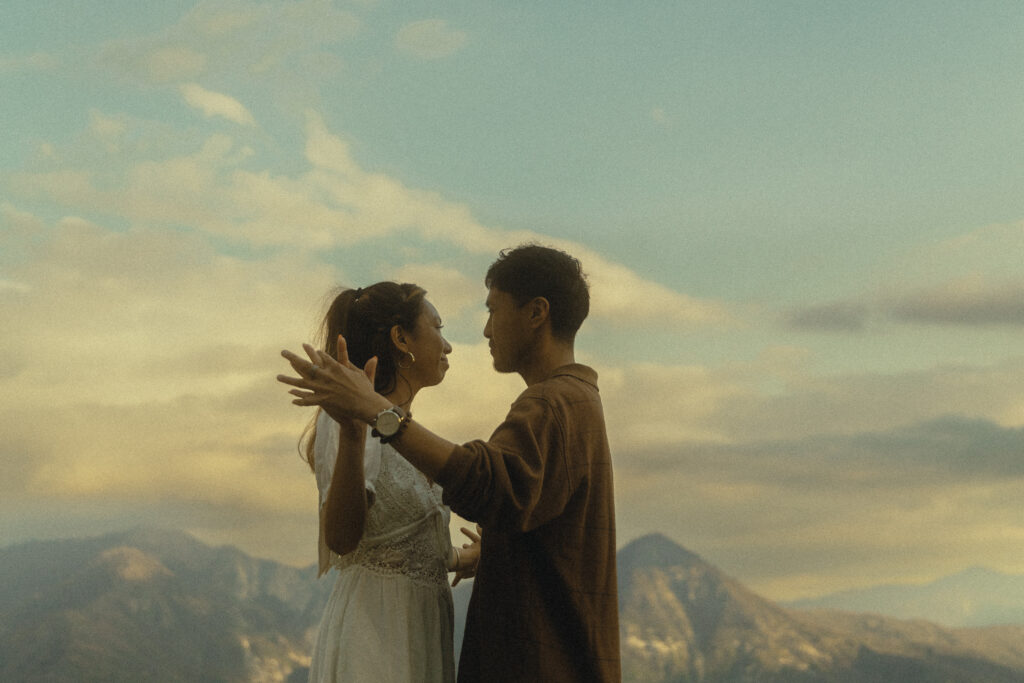 a couple stand facing each other clasping their hands together with mountains and the soft beginnings of a sunset behind them