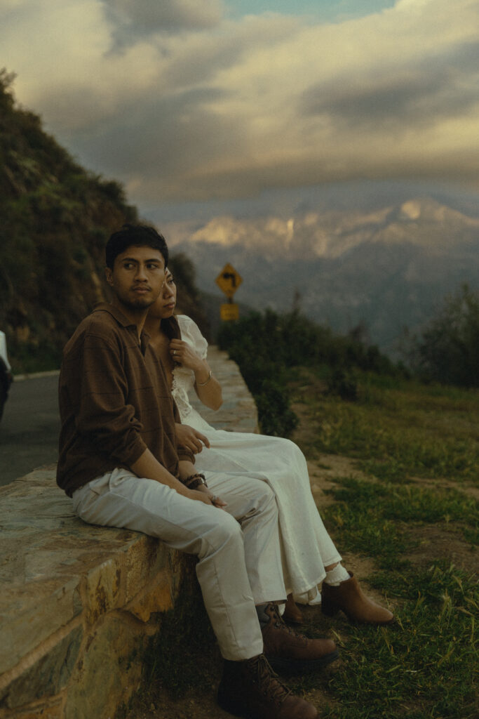 a man sits next to his fiancee on a stone bench and looks to the side in front of a mountain view