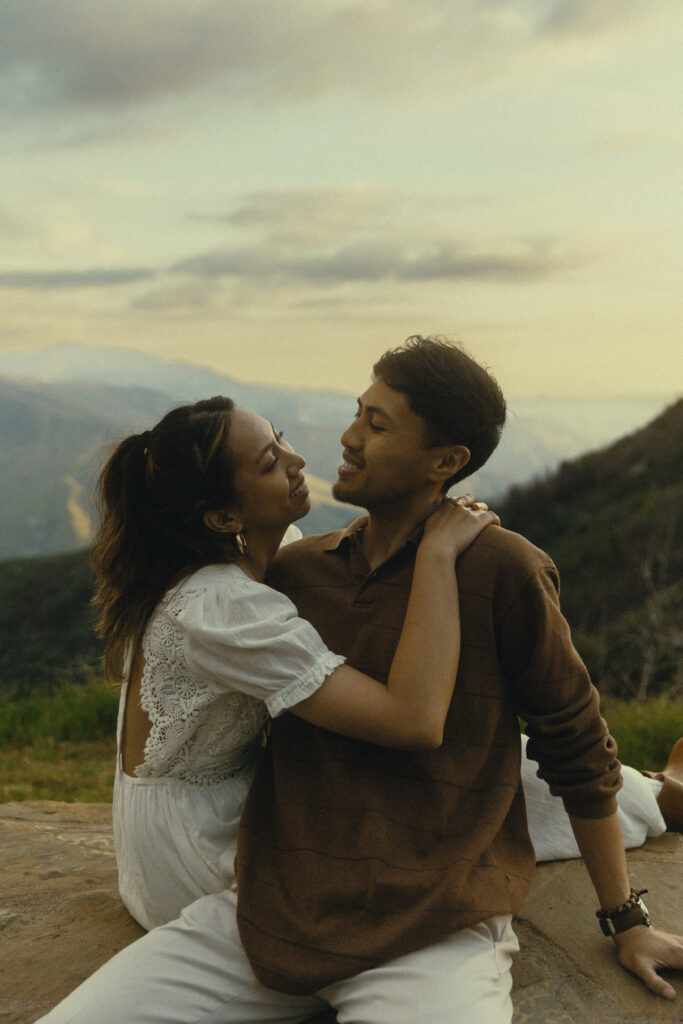 a couple smiles as they look at each other, she sits behind him with an arm around his chest as he leans on his arm on the other side of her with mountains and a soft sky behind them