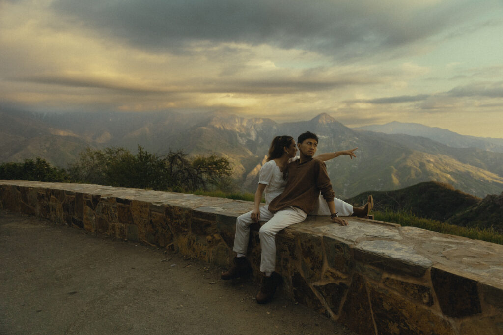 a woman points to something in the distance as she sits next to her fiancee facing the other direction with the mountains behind him