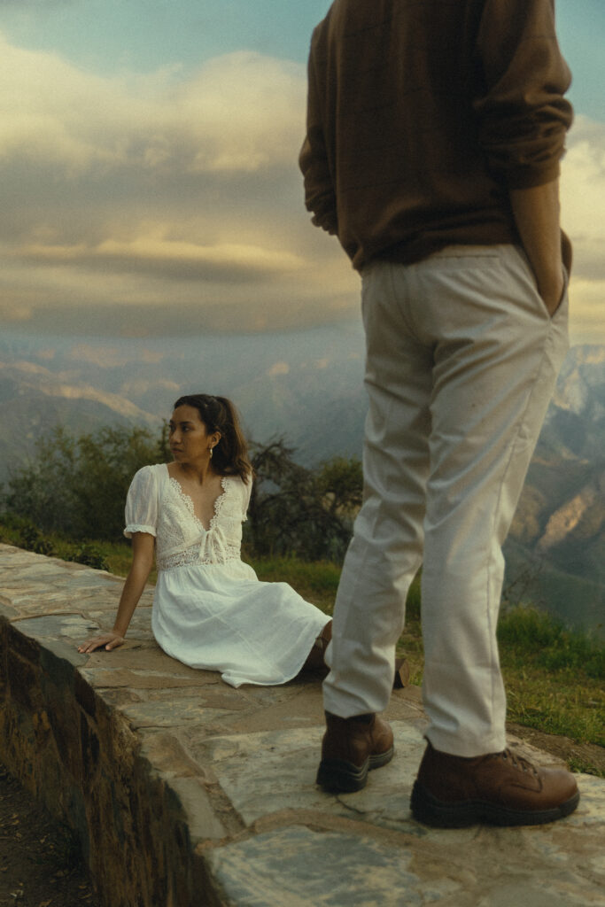 a man stands on a stone bench with his fiancee laying next to him as they look out at the mountains of sequoia national park engagement photos
