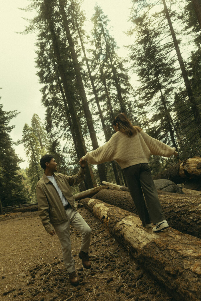 a man holds his fiancee's hand as she holds her arms out for balance as she walks across fallen trees