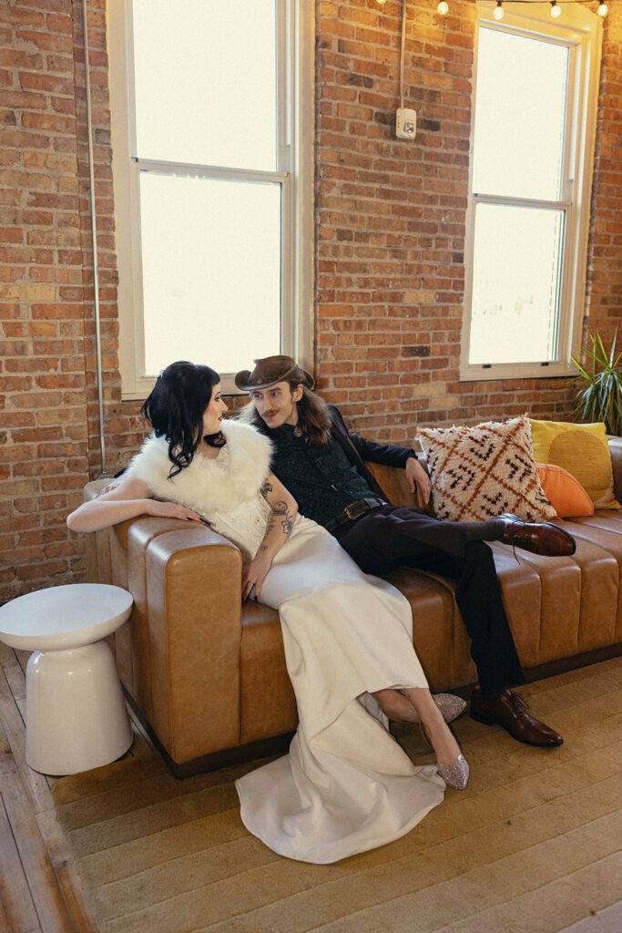 A Michigan newlywed couple poses together in their wedding attire on a brown leather couch at Record Box Loft, a venue for weddings southwest michigan