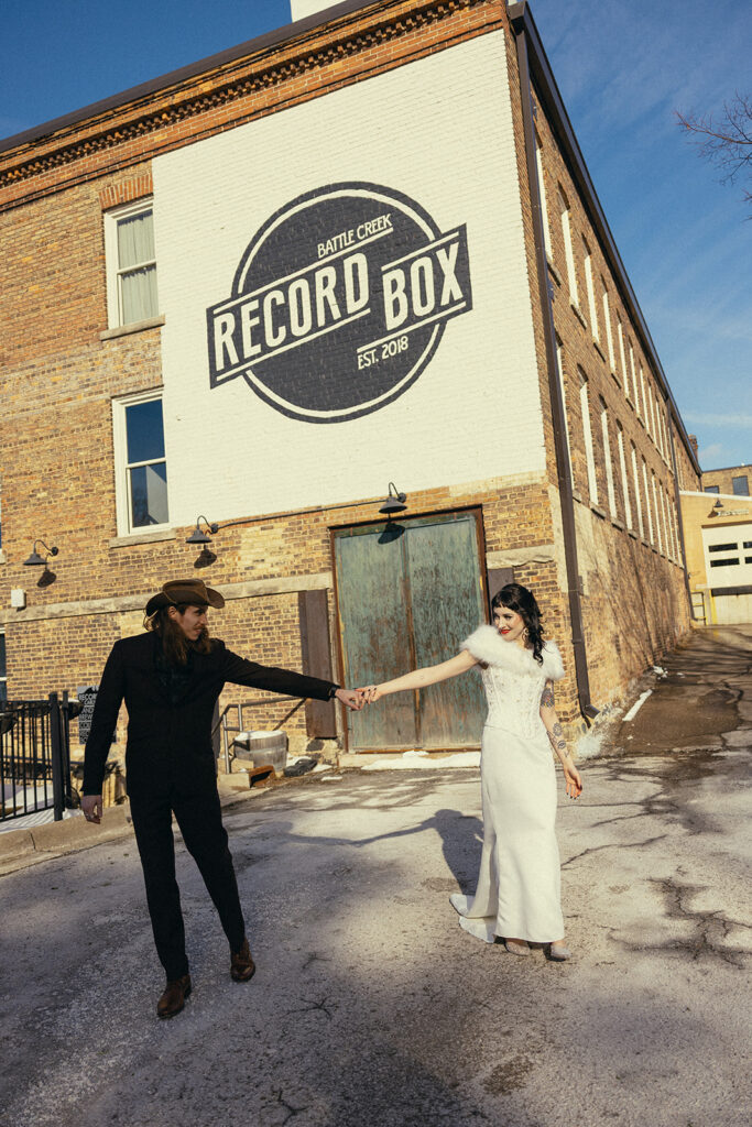 A Michigan couple wearing Western 70s wedding attire hold hands outside Record Box Loft, their Battle Creek wedding venue