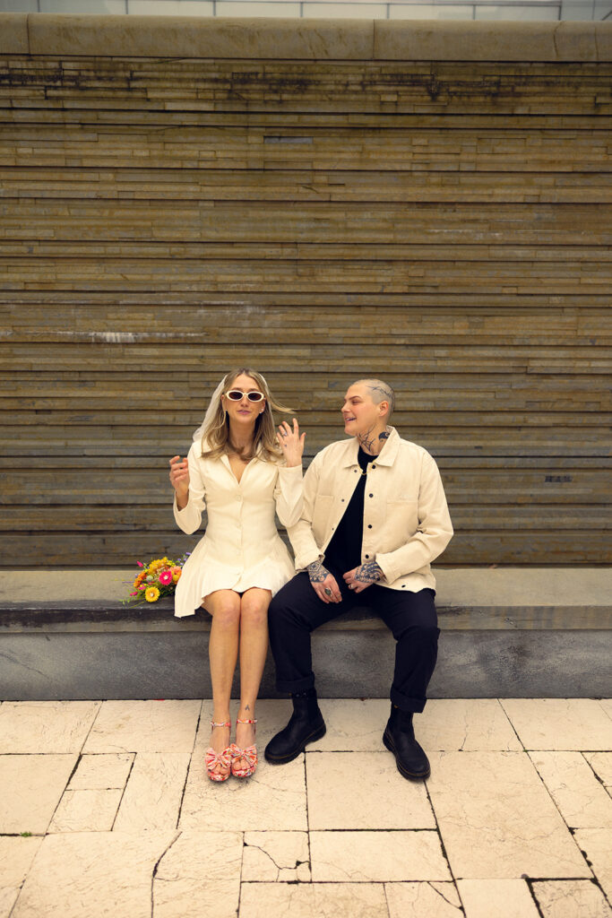 A Bride wearing a short white dress & white sunnies adjusts her veil as her spouse looks on with a smile before their courthouse wedding grand rapids mi