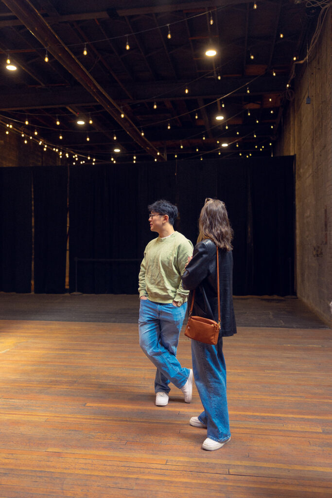 A Michigan couple stands underneath string lights at The Jam Handy, a Detroit wedding venue, during a photoshoot with Michigan engagement photographer Anika B Photography