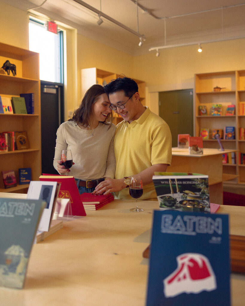 A couple smiles as they enjoy a glass of wine while browsing books during their photoshoot by a Detroit engagement photographer 