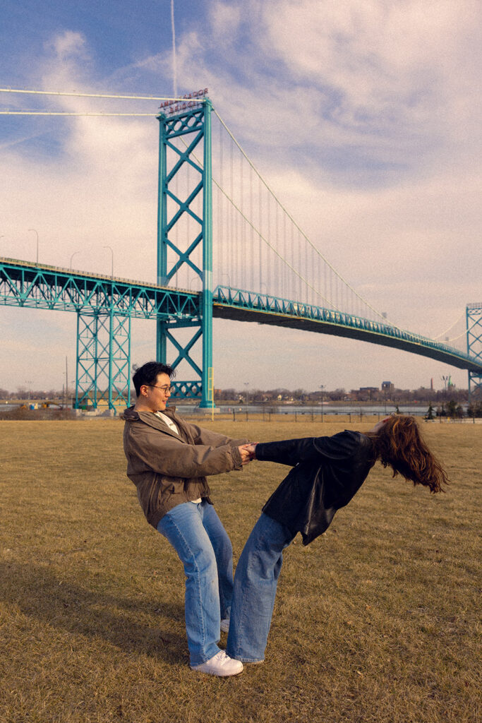 A couple holds hands in the grass near the Ambassador Bridge, one of the outdoor options for Detroit engagement photo locations