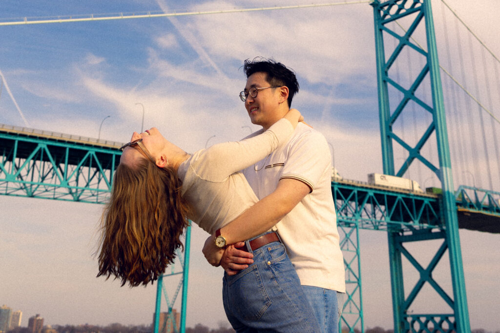 A Michigan couple wearing jeans & white shirts laugh and embrace beneath a blue sky near the River Walk during their Detroit engagement photos 