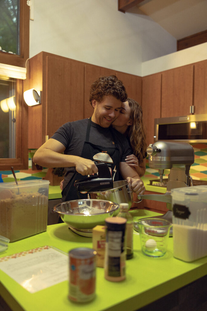 A brunette woman kisses her fiancé on the cheek while he mixes cookies in the kitchen of their MCM home during Michigan engagement photos 
