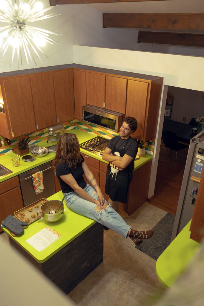 A young couple chat leaned against their green countertops of their MCM kitchen during their 