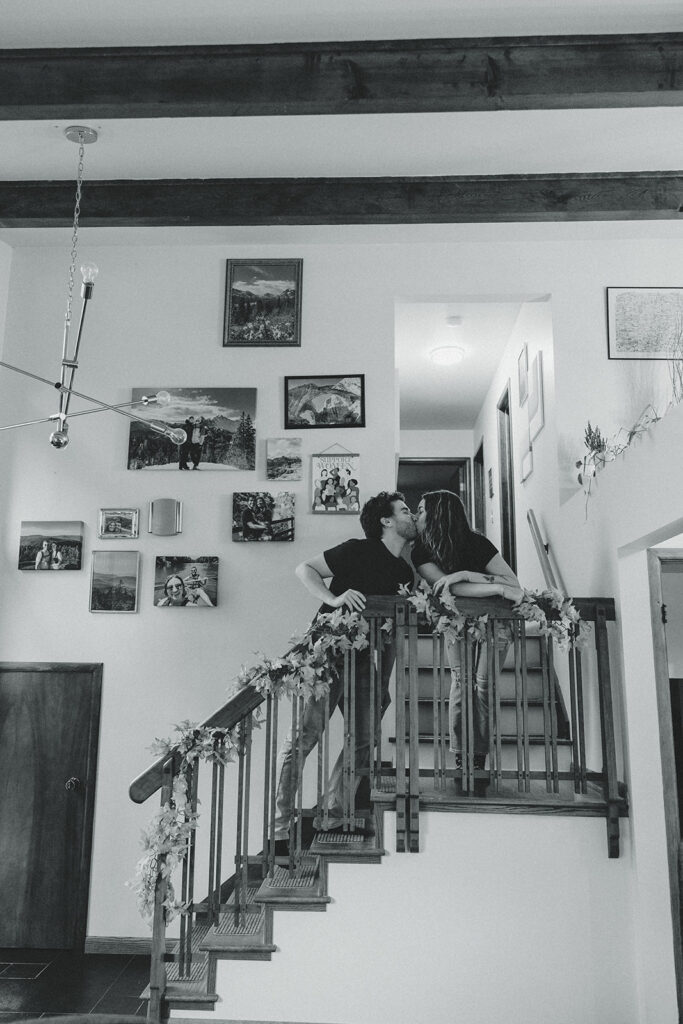 A black and white image of a couple sharing a kiss on the staircase of their vintage house in michigan during an in-home engagement session with Anika B Photography 