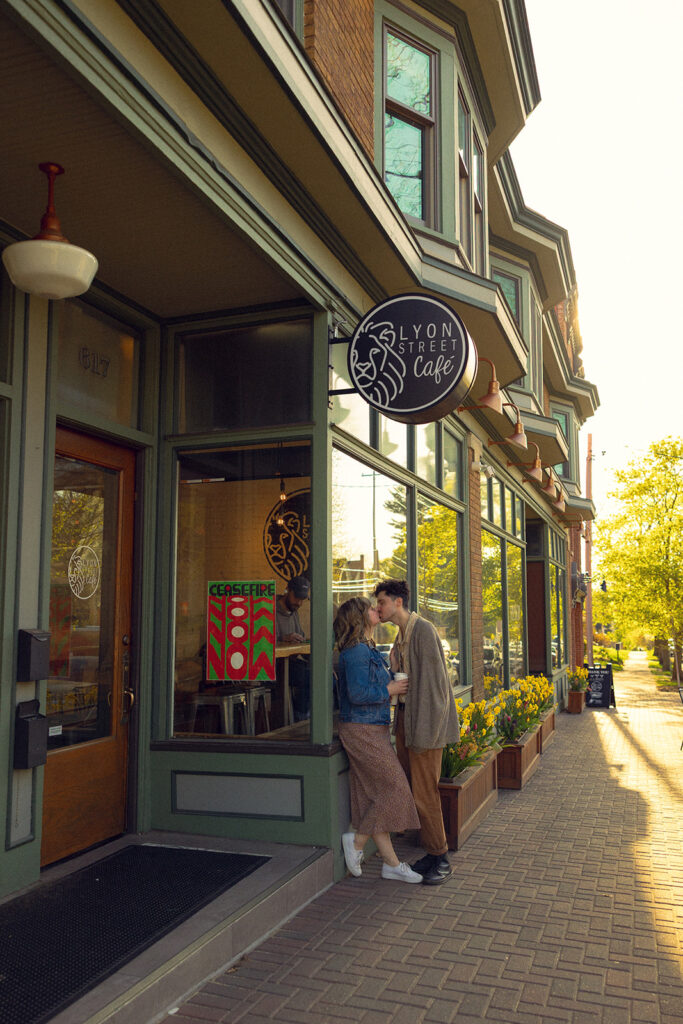 A couple share a kiss outside Lyon Street Cafe during engagement photos in MI