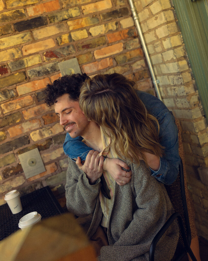 A woman wearing a denim jacket kisses her fiancé on the cheek outside Lyon Street Cafe during engagement photography in Grand Rapids MI