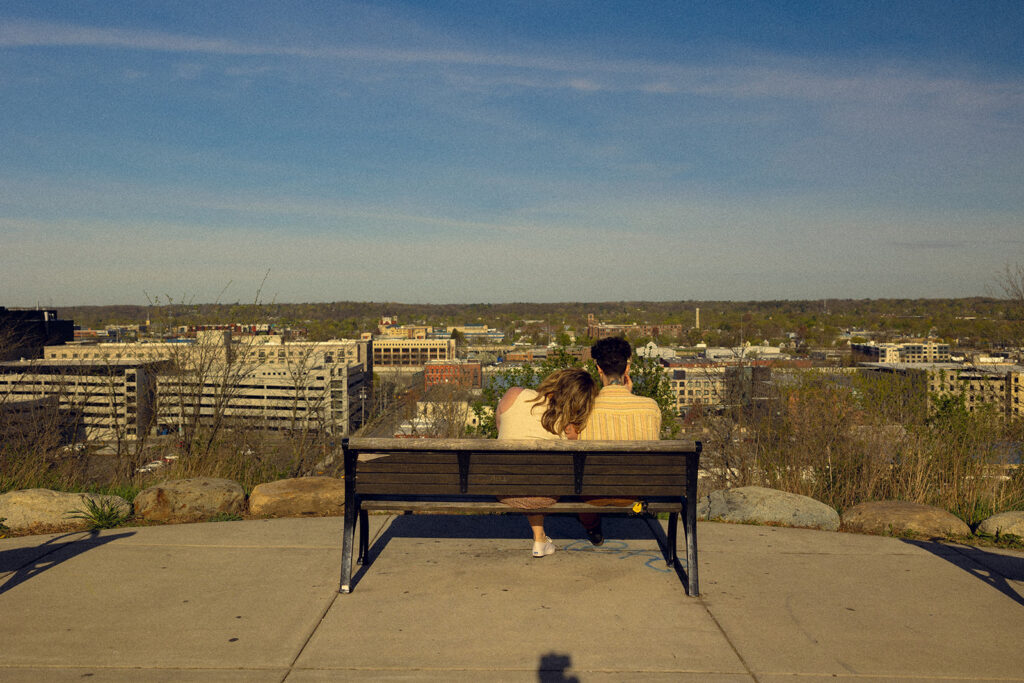 A couple is seated on a bench at Lookout Park overlooking the city during Michigan engagement photos