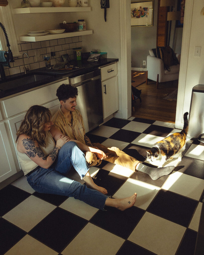 A couple are seated on the b+w checkered floor if their Grand Rapids home as they play with their calico cat during engagement photography Grand Rapids MI