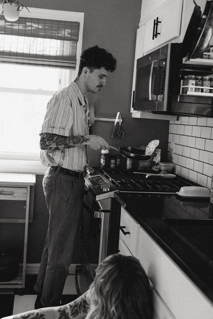 A b+w image of a man preparing eggs in the kitchen of the home he shares with his fiancée during engagement photos Michigan
