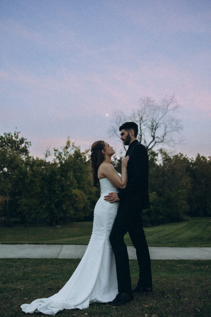 A bride in a white gown embraces her groom in a black suit at sunset following their Michigan wedding venue