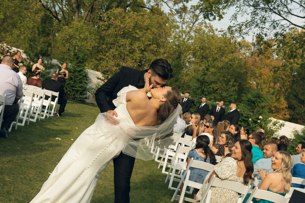 A bride and groom share a kiss at the end of the aisle at one of the outdoor Michigan wedding venues 