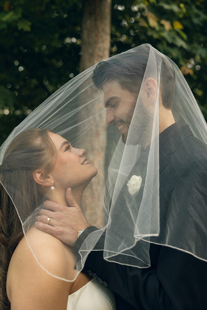Newlyweds smile at each other from beneath the bride's veil in a bridal portrait taken by West Michigan wedding photographer