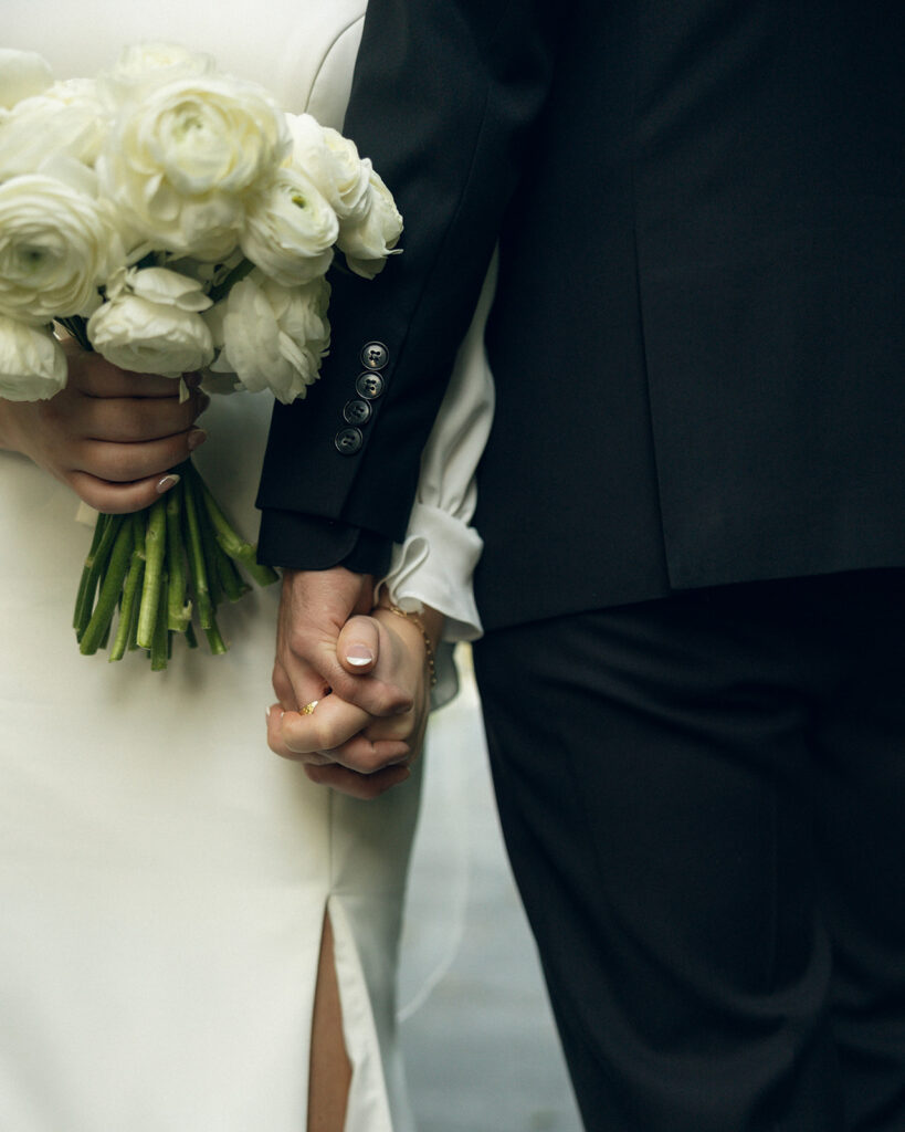 A close up photo of newlyweds holding hands as the bride also holds her bouquet of white florals at their outdoor wedding in west michigan
