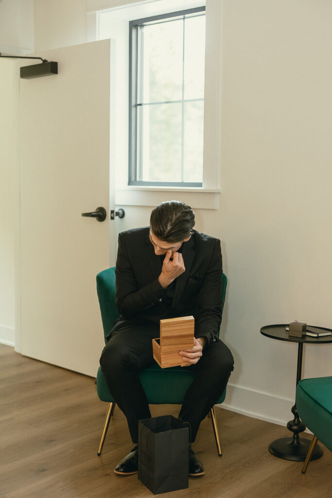 A groom in an all black suit gets emotional as he opens a gift from his bride at one of the more unique wedding locations in michigan