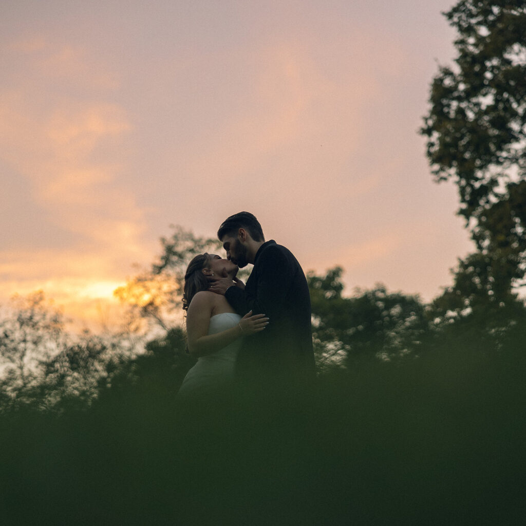 Newlyweds share a kiss before a coral and purple sky at one of outdoor michigan wedding venues