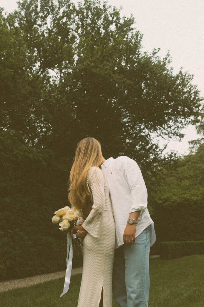 An editorial engagement photo depicts a young woman holding a bouquet of flowers behind her back as her partner leans in for a kiss at the Ford House in grosse Pointe MI 