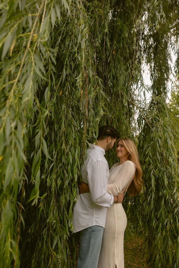 A candid engagement photo focuses on a young couple smiling amongst the landscaping at the henry ford wedding venue