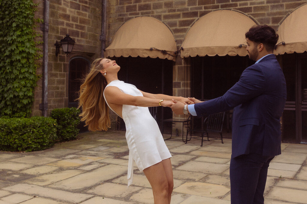 A man wearing a navy suit twirls his fiancée wearing a short, white dress during their Ford Mansion Grosse Pointe engagement photos