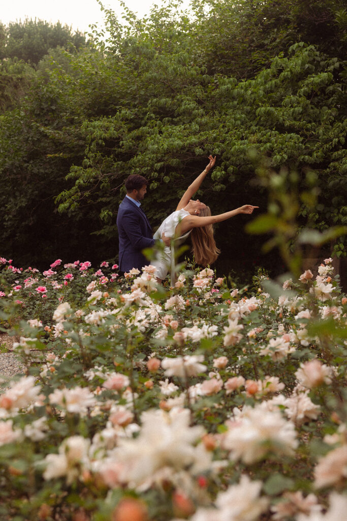 A man in a navy suit dips his soon-to-be-bride amongst the gardens at the henry ford wedding venue during their Grosse Pointe engagement photos 