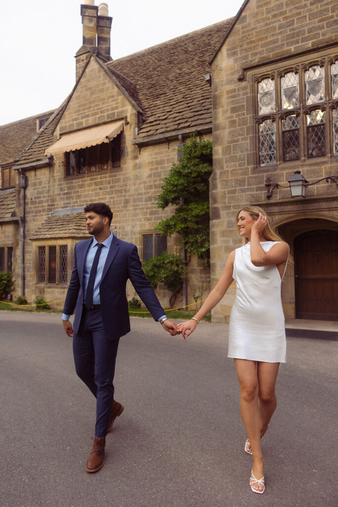 A soon-to-be-wed couple wearing  a navy suit and a short white dress hold hands during engagement photos taken during a Ford House Tour