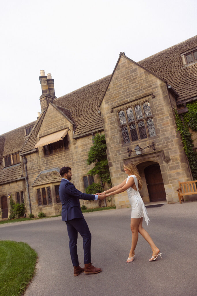 An engaged couple holds hands and twirls in the drive during a Ford House Tour engagement shoot with Anika B Photography 