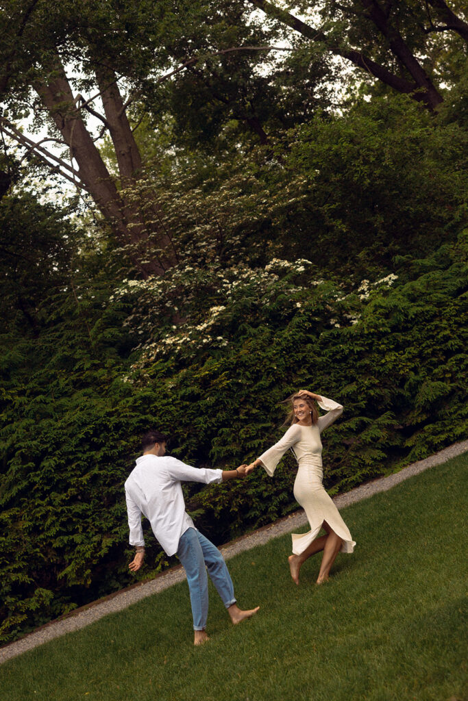 An editorial engagement photo shows a young couple holding hands and running through the grass of the estate during a Ford House tour 