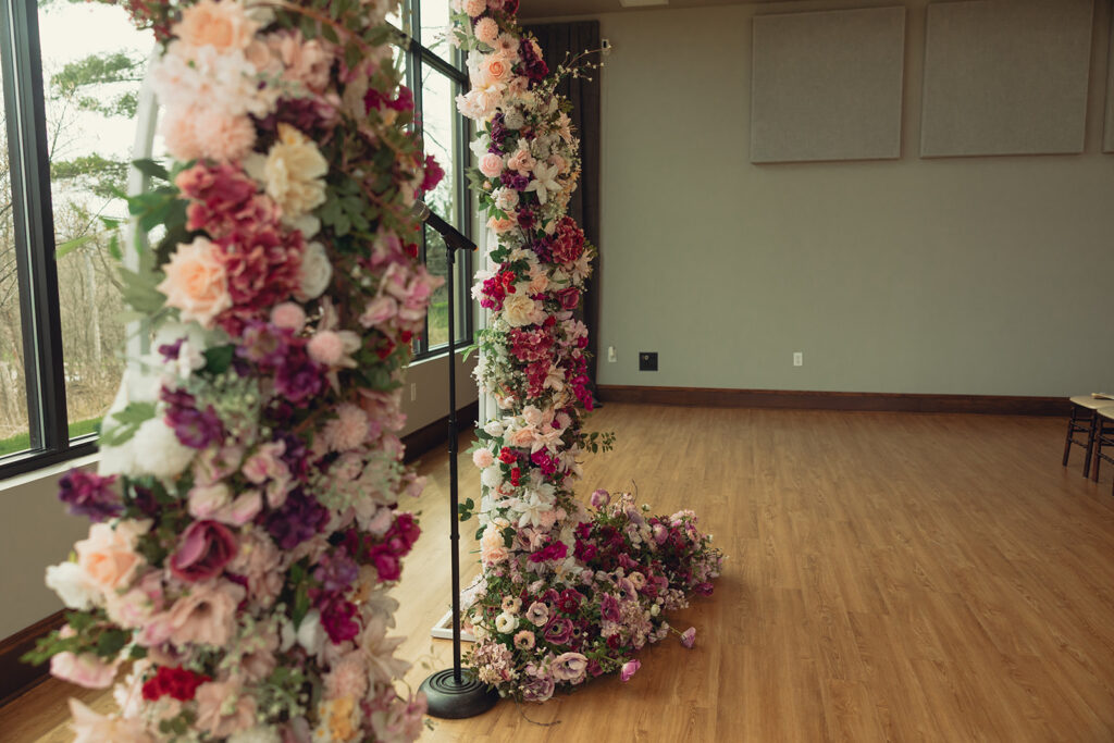 A floral covered arch before a large window is photographed at a Venue3Two wedding