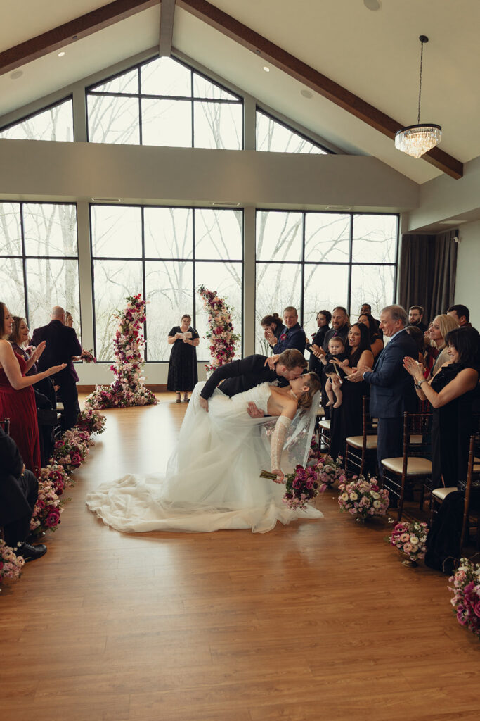 A groom in a black tux dips his new wife for a kiss at the end of the aisle following their wedding ceremony grand rapids mi