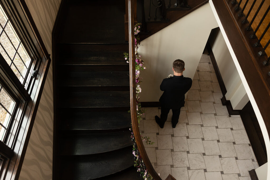 A groom awaits a first look at his bride at the bottom of an ornate staircase at Venue3Two in Grand Rapids, MI