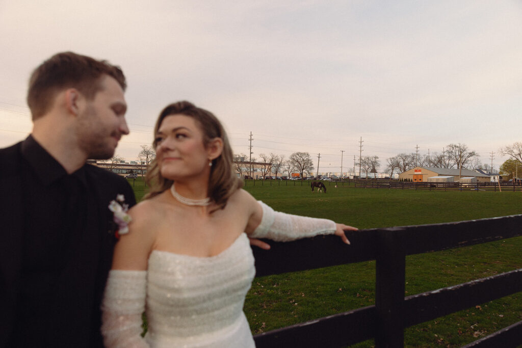Newlyweds smile as they lean against a fence at sunset with horses in the background at Venue3Two