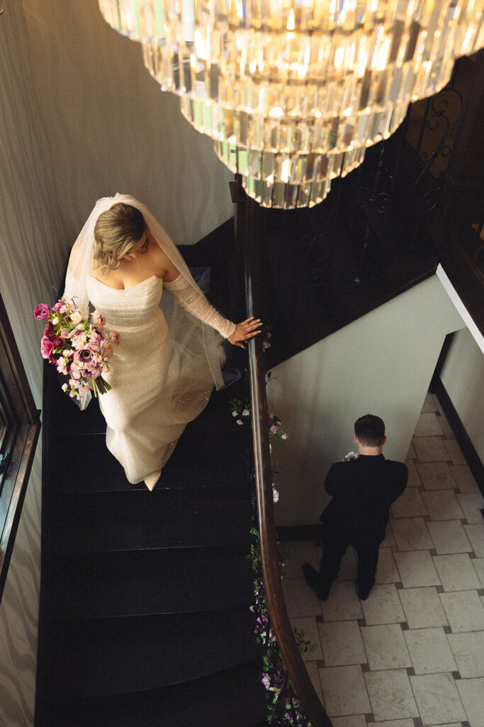 A bride wearing her gown & veil walks down a staircase beneath a crystal chandelier to her awaiting groom at Venue3Two, an all inclusive wedding venues in michigan